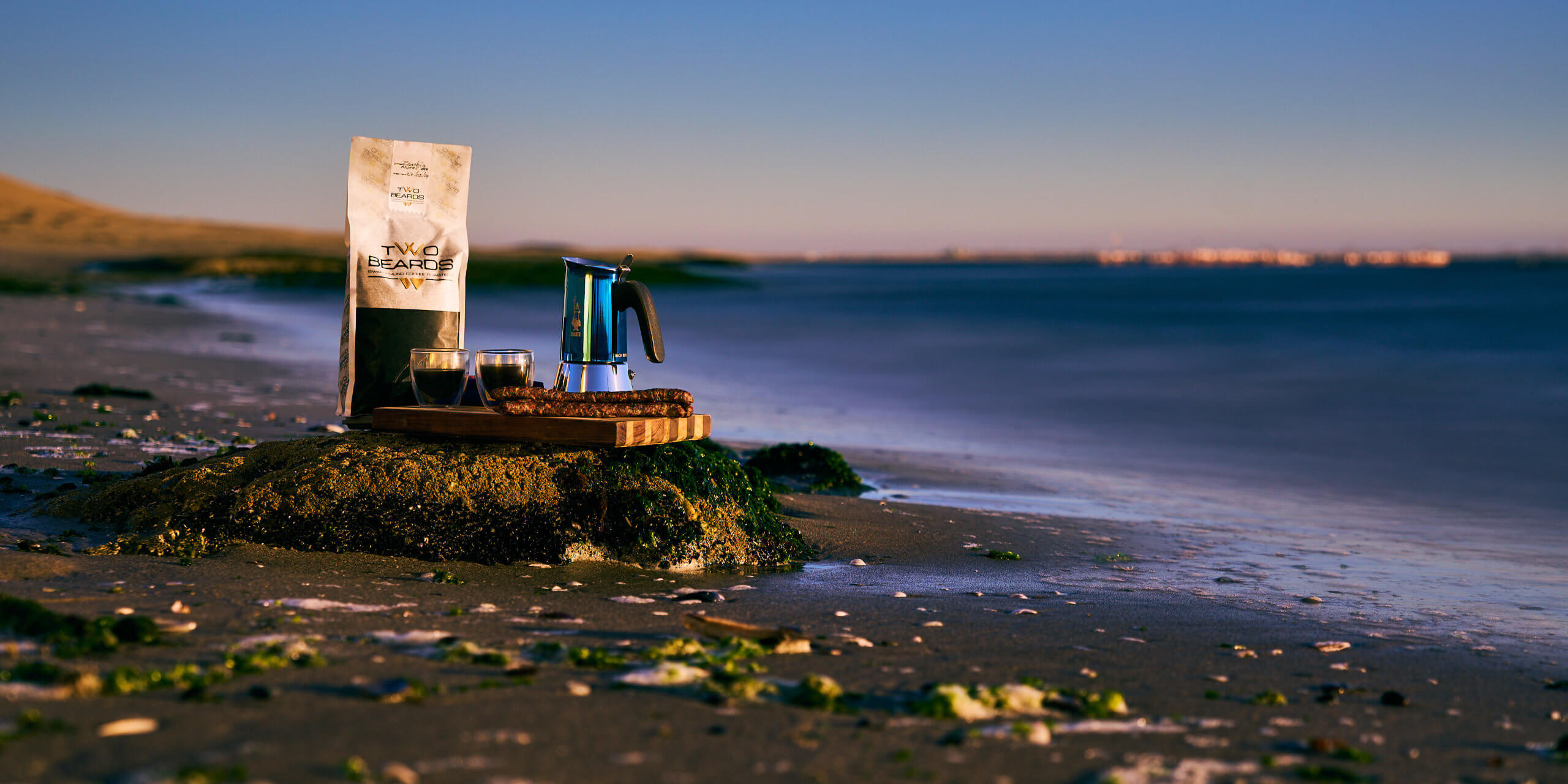A tranquil beach scene at dusk featuring a bag of Two Beards Coffee, a metallic coffee maker, and two cups on a wooden board atop a seaweed-covered rock, with the ocean and distant flamingos in the  background.