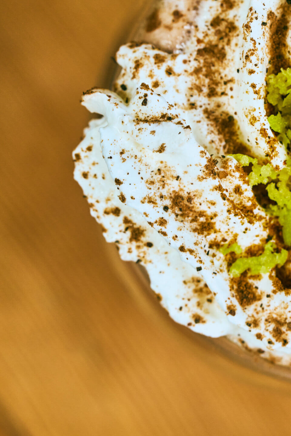 A coffee featuring fluffy white whipped cream sprinkled with brown spices, with a layer of green crumbles beneath, all presented on a beige surface.